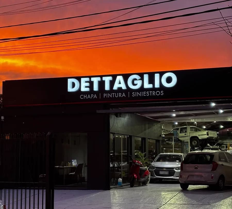 Frente de la sucursal de Malvín del taller Dettaglio al atardecer, con el cielo naranja: el cartel negro iluminado con el nombre y el portón abierto con los autos adentro