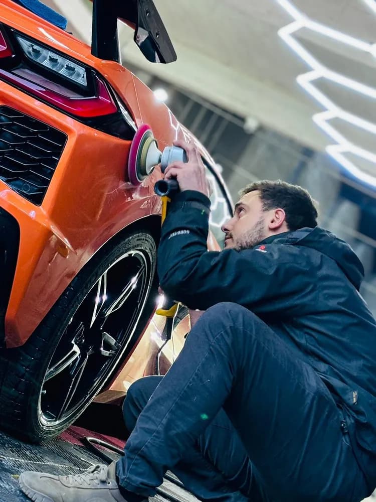 Técnico del taller puliendo con rotativa el frente de un deportivo naranja, bajo las luces hexagonales
