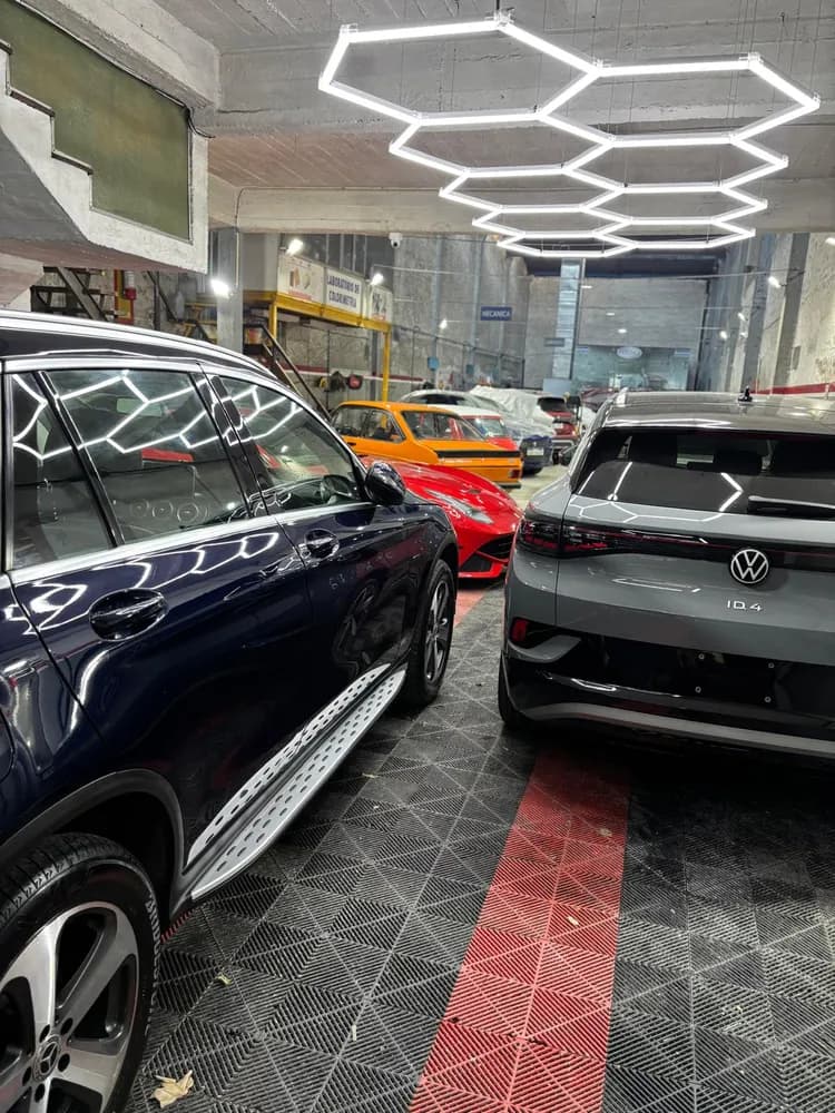Varios autos alineados dentro del taller, con la lámpara hexagonal reflejada sobre la chapa